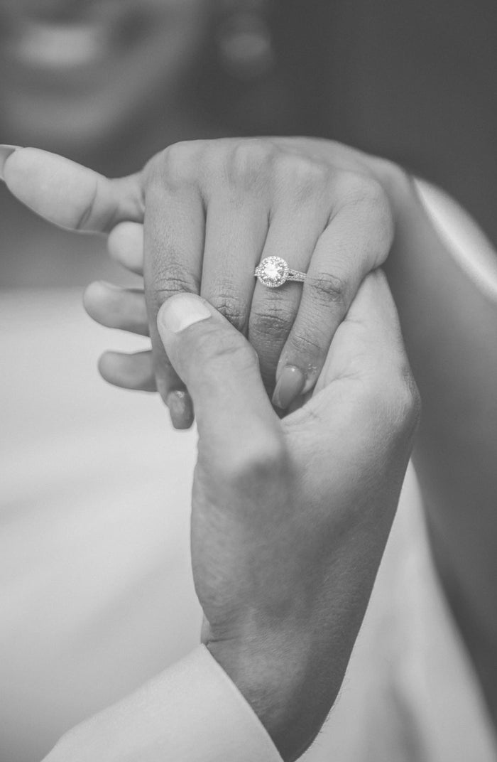 A close-up black and white photograph of a ring being placed on a finger, with two hands visible and soft blurred clothing in the background.